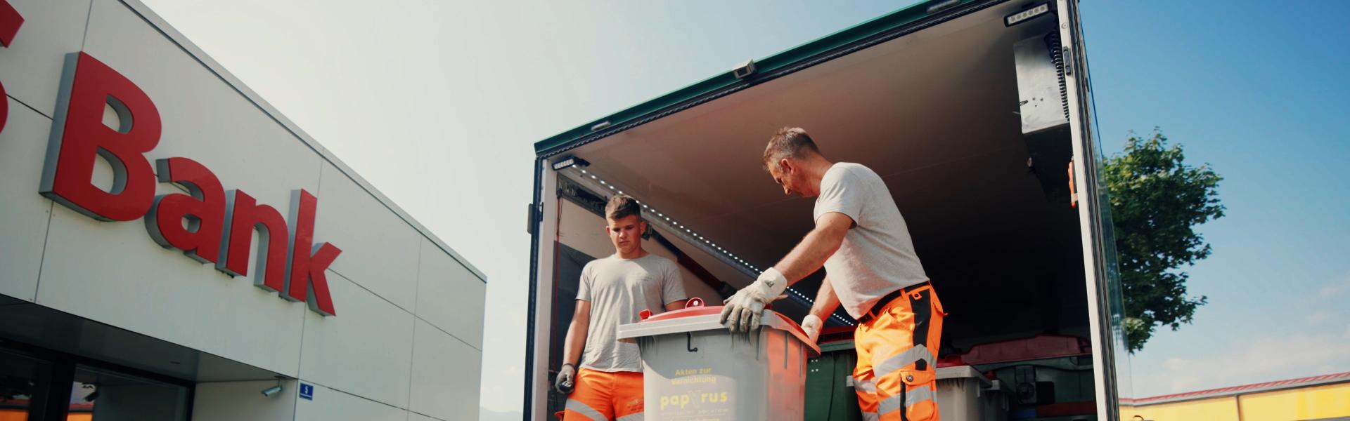 Zwei Mitarbeiter verladen einen 240 l Behälter mit Akten in den Kastenwagen